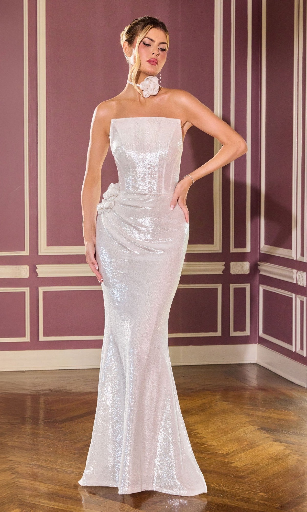 A woman poses indoors against a mauve wall with white trim, wearing the off white Ladivine J905 long prom dress, which is strapless and sequined, featuring floral accents at the neck and hip.